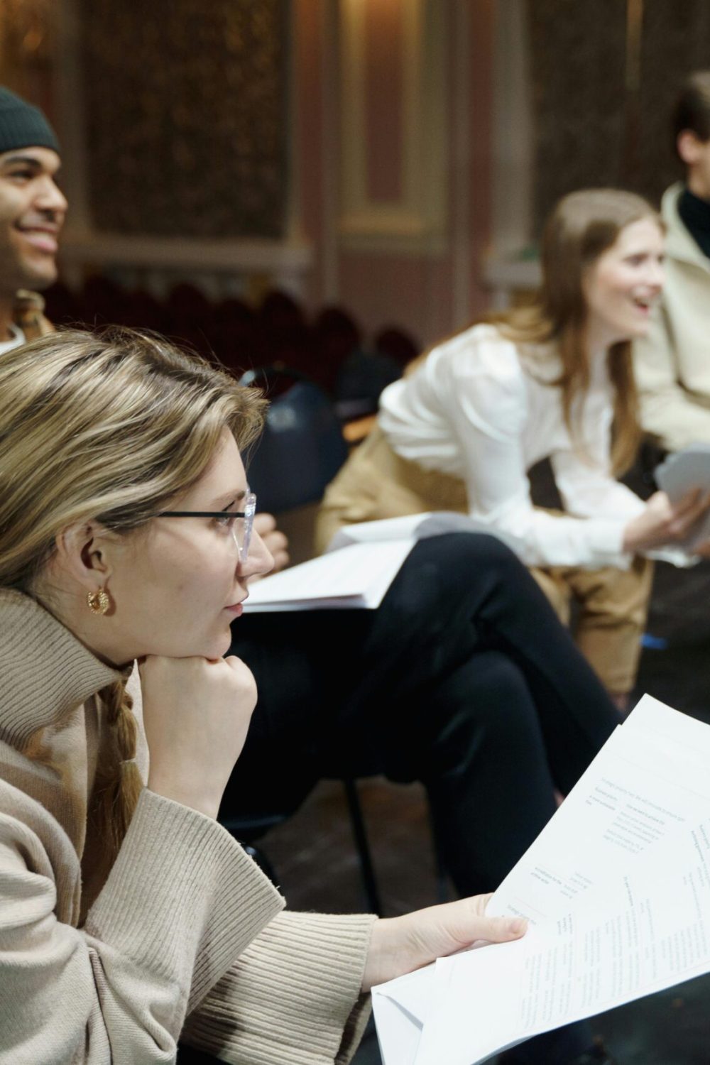 Actors rehearsing a play on stage, holding scripts and discussing roles in a theater setting.